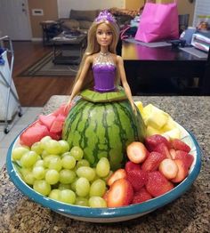 a doll sitting on top of a watermelon bowl filled with grapes and strawberries