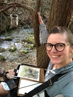 a woman with glasses holding an open book in front of a tree and water stream