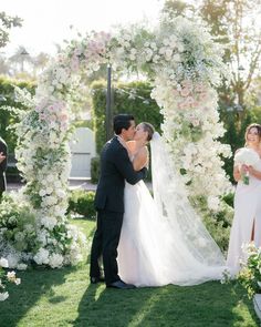 a bride and groom kissing in front of an archway with flowers on the grass at their wedding