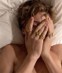 a shirtless man covers his face while laying in bed with two gold rings on his hands