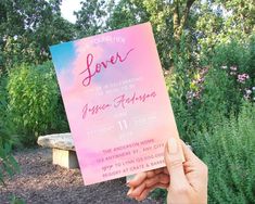 a person holding up a pink and blue wedding card in front of some trees, bushes and flowers