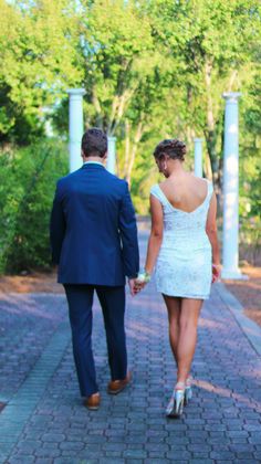 a man and woman walking down a sidewalk holding hands, dressed in blue suit and white dress