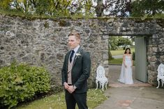 a man standing in front of a stone wall next to a woman wearing a wedding dress