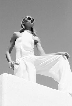 a woman standing on top of a white wall with her hands in her pockets and wearing sunglasses