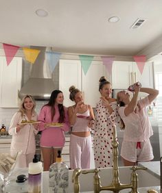 four women standing in a kitchen taking pictures with their cell phones and drinking from the faucet