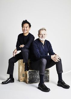 two men sitting on top of suitcases posing for a photo in front of a white background