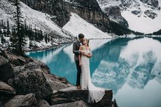 a bride and groom standing on the edge of a mountain overlooking a lake with snow covered mountains in the background