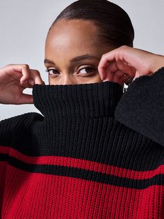 A person with dark, pulled-back hair is partially covering their face with the high collar of the Stay Cozy Sweater Black Red Stripe by Sanctuary Clothing. They are looking directly at the camera, with hands raised to adjust the collar. The background is plain and light-colored. Black Funnel Neck Sweater For Layering, Winter High Neck Black Tops, Black Cozy Funnel Neck Top, Cozy Black Funnel Neck Top, Cozy Stretch Black Tops, Winter Black Top With Ribbed Cuffs, Fitted Black Sweater With Funnel Neck, Cozy Black Top For Streetwear, Trendy Black Tops For Cold Weather