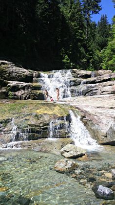 two people are playing in the water near some rocks and trees, while another person is standing at the base of a waterfall