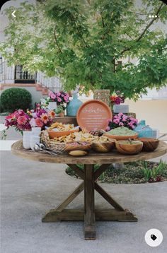 a wooden table topped with lots of food next to a lush green leafy tree