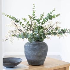 a vase filled with lots of green plants on top of a wooden table next to a bowl