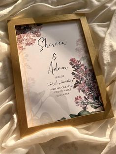 a wedding card with flowers on it sitting on a white cloth covered bed sheet in front of a wooden frame