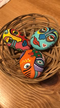 three painted rocks sitting on top of a wooden table next to a wire basket filled with eggs