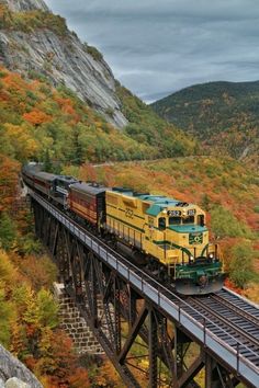 a train traveling over a bridge surrounded by mountains and trees in the fall time with colorful foliage