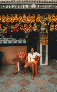 a woman sitting in front of a fruit stand with oranges hanging from the ceiling