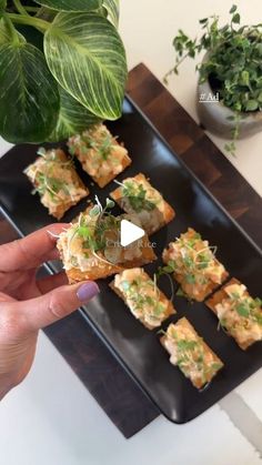 someone is holding their finger up to some food on a black tray with green plants in the background