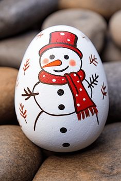 an egg with a snowman painted on it is sitting on some rocks and has a red scarf around its neck