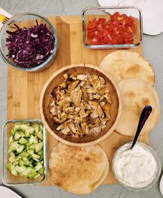 a wooden cutting board topped with bowls filled with food next to sliced vegetables and pita bread