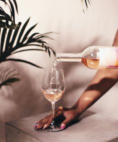 a woman pouring wine into a glass on top of a wooden table next to a potted plant