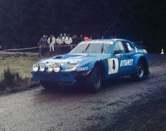 a blue race car driving down a dirt road with people watching from the sidelines