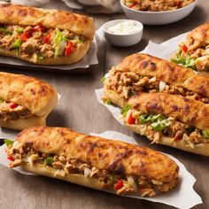 several sandwiches with meat and vegetables on paper lined up next to bowls of dips