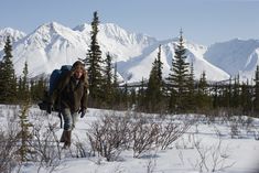 a woman with a backpack walking through the snow in front of some trees and mountains