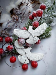 two white doves with red berries and pine needles on the snow covered ground next to evergreen branches