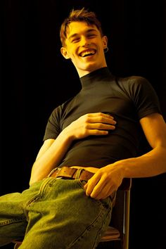 a smiling man sitting in a chair with his hands on his hips and wearing green jeans