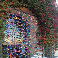 colorful paper cranes hanging from the side of a building next to trees and flowers in front of it