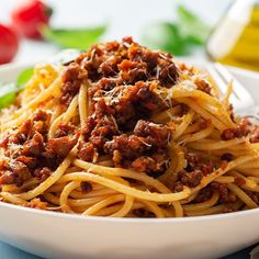 a white bowl filled with spaghetti and meat on top of a blue table cloth next to tomatoes