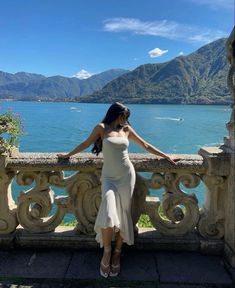 a woman in a white dress standing on a balcony looking out at the water and mountains