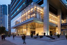 people are walking around in front of a building that has glass windows and lights on it