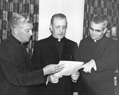 black and white photo of three men standing in front of a wall with books on it