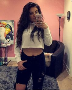 a woman taking a selfie with her cell phone in front of the camera, wearing ripped jeans and a white crop top