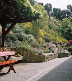 a wooden bench sitting on top of a sidewalk next to a lush green hillside covered in flowers