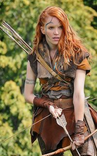 a woman with red hair is dressed in costume and holding two arrows while walking through the woods