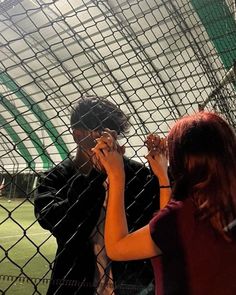 two people standing behind a chain link fence on a soccer field, one holding something up to his face