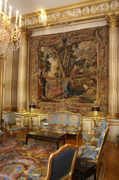 a large tapestry hanging in the middle of a living room with blue chairs and chandeliers