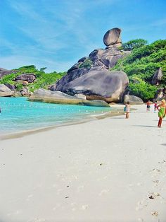 people are walking on the beach near large rocks and clear blue water, surrounded by green trees