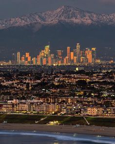 the city skyline is lit up at night with snow capped mountains in the background