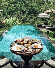 an outdoor table with food and drinks near a swimming pool in the middle of a garden