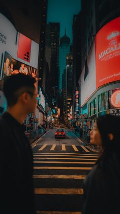 a man and woman standing in the middle of a crosswalk at night with city lights behind them