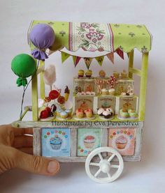 a hand holding up a small toy cart with cupcakes and balloons on it