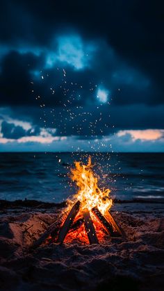 a campfire on the beach with dark clouds in the sky and water behind it