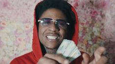 a man with glasses holding money in front of his face and wearing a red hoodie