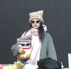 a woman holding a stuffed animal wearing sunglasses and a knitted hat is standing in front of a table