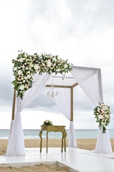 an outdoor wedding set up with white drapes and flowers on the side of the beach