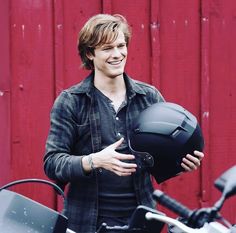 a young man smiles while holding his motorcycle helmet in front of a red barn door