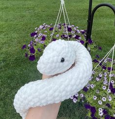 a hand holding a white knitted object in front of purple flowers and a black pole