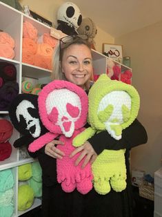 a woman holding two crocheted stuffed animals in front of a wall of skulls